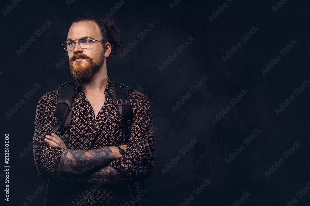 Portrait of a redhead hipster student in glasses dressed in a brown shirt, holds a backpack, posing at a studio. Isolated on the dark background.