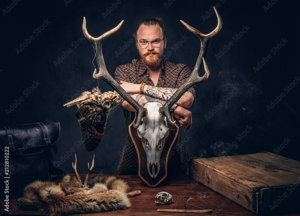 Φωτογραφία Redhead taxidermist hipster male in sunglasses dressed in a brown shirt, standing near a table with handmade trophy, owl scarecrow, and the fox skin