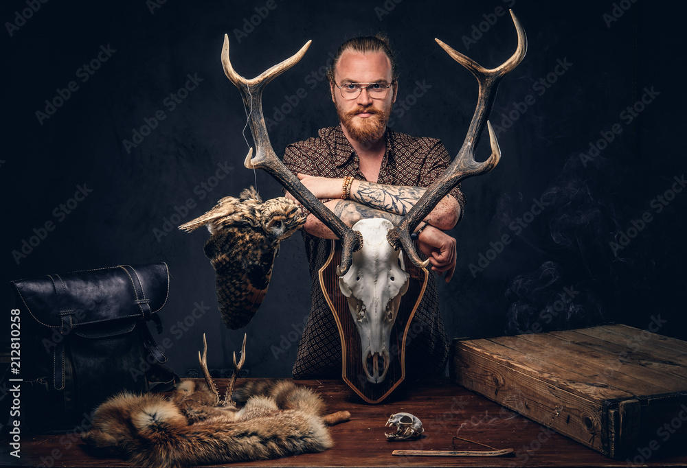 Φωτογραφία Redhead taxidermist hipster male in sunglasses dressed in a brown shirt, standing near a table with handmade trophy, owl scarecrow, and the fox skin