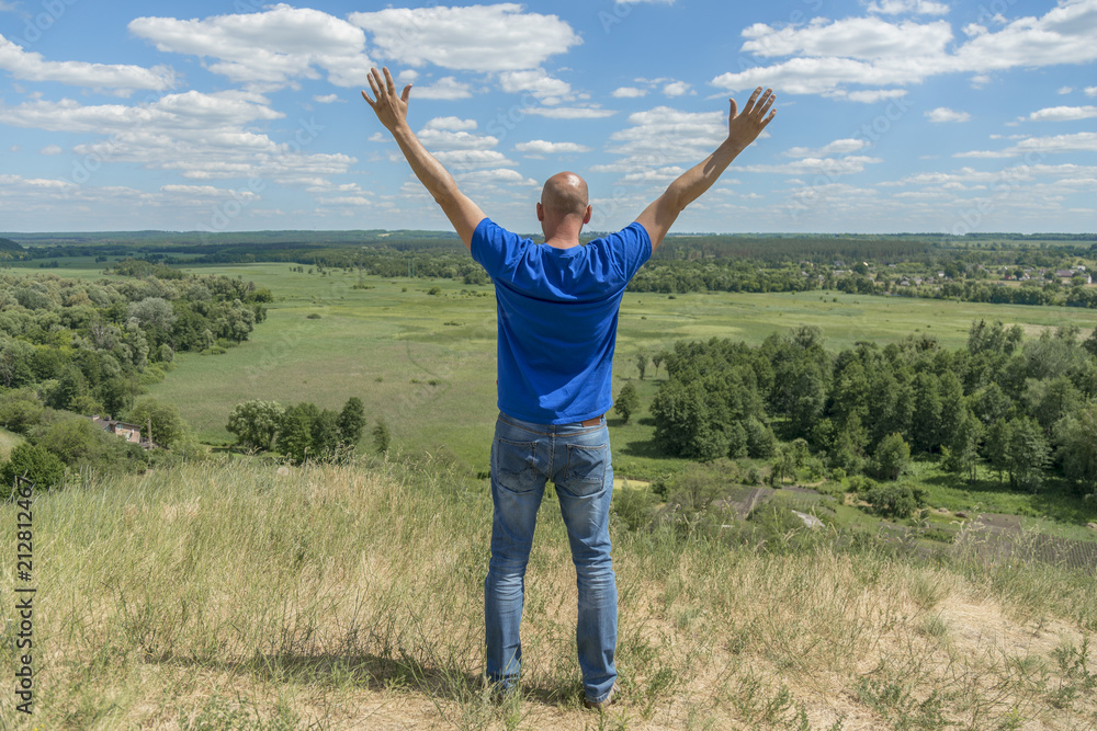 Young man is travelling in summer holidays. man in modern dress is standing on the hill and looking at the field. Blue sky and white clouds. Freedom and lifestyle concept. Beautiful nature Young man is travelling in summer holidays. man in modern dress is standing on the hill and looking at the field. Blue sky and white clouds. Freedom and lifestyle concept. Beautiful nature