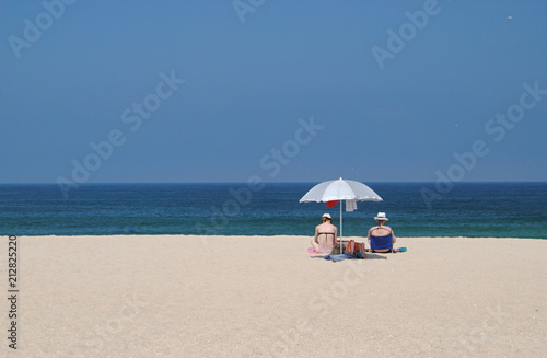 Duas mulheres sentadas na areia da praia perto do mar com o guarda-sol aberto a apreciar o horizonte na praia