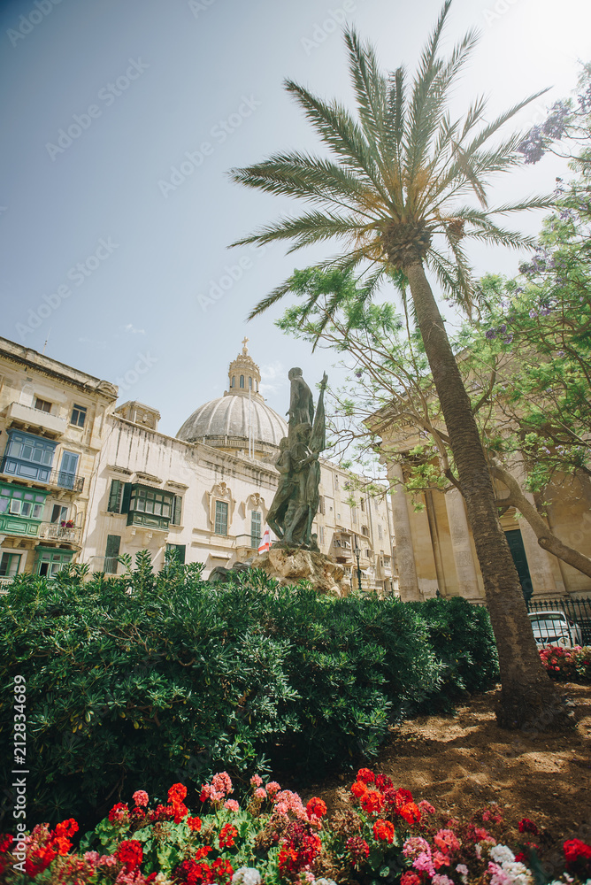 Beautiful view from square with green bushes. flowers and palm of satue of saint and cathedral dome, church in Valetta, Malta