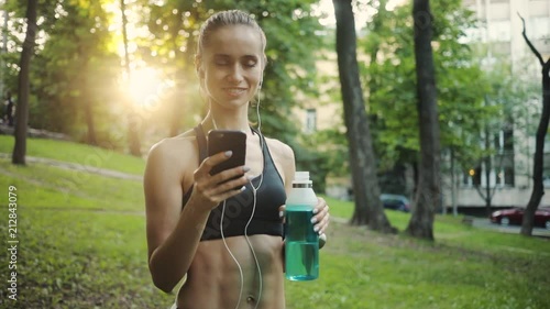 Happy young woman use smartphone and headphone drink water in park sun outdoor health hand girl tree sport fitness summer nature internet technology runner exercise phone workout mobile cellphone