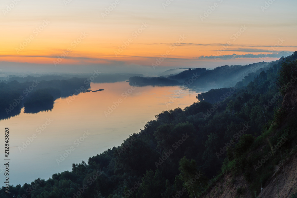View from high shore on broad river. Riverbank with forest under thick fog. Dawn reflected in water. Yellow glow in picturesque predawn sky. Colorful morning atmospheric landscape of majestic nature.