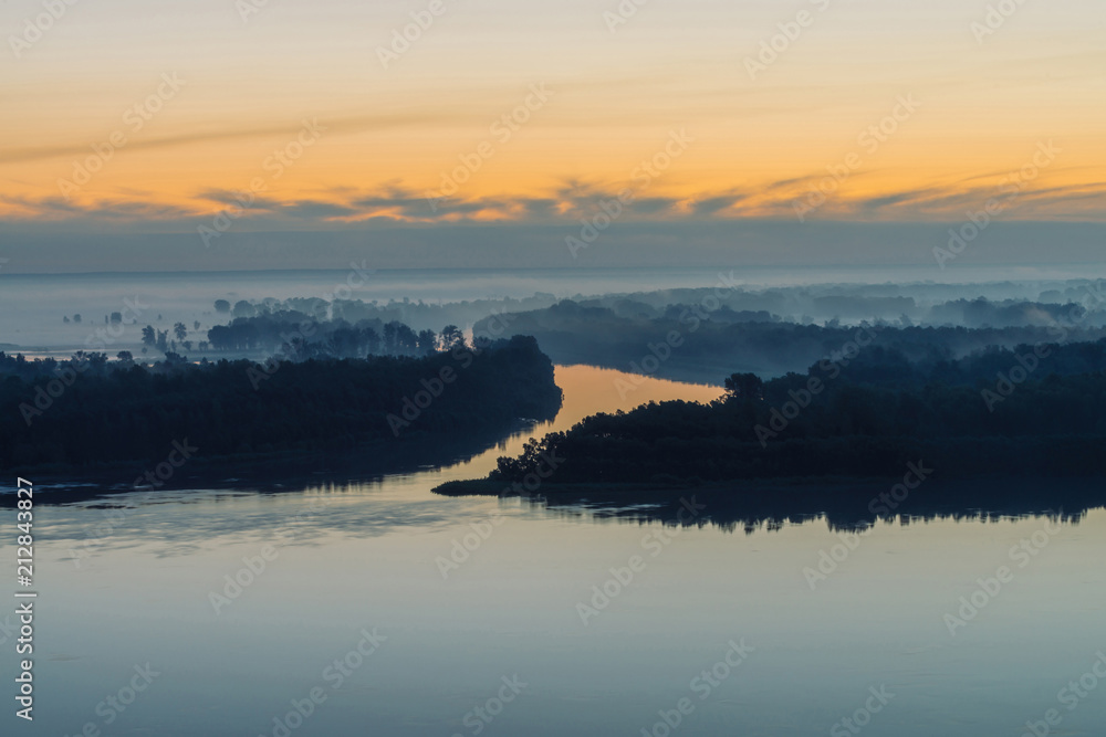 Broad river flows along coast with forest under thick fog. Early blue sky reflected in water. Yellow stripe in picturesque predawn sky. Colorful morning atmospheric landscape of majestic nature.