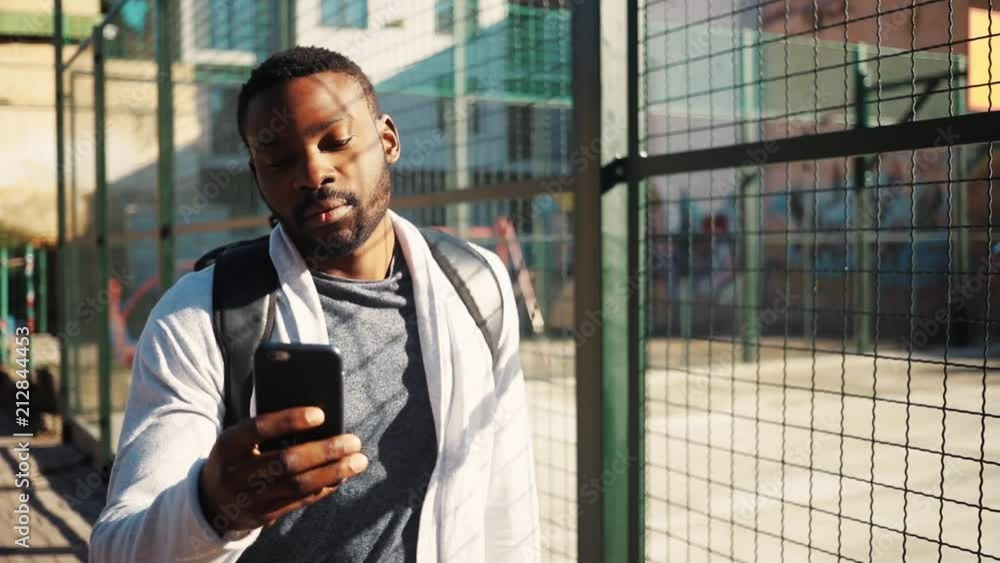 Good-looking student getting mobile phone from pocket. Cute surprised young African man looking at screen of smartphone, laughing. Street. Outdoors. Good-looking student getting mobile phone from pocket. Cute surprised young African man looking at screen of smartphone, laughing. Street. Outdoors.