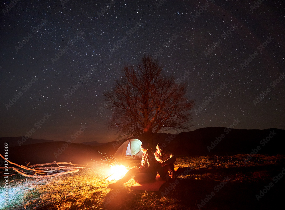Camping night in mountains. Silhouettes of couple tourists sitting, man and woman lit by burning campfire under starry sky. Tent, big tree and distant hills on background. Tourism and travel concept - obrazy, fototapety, plakaty 