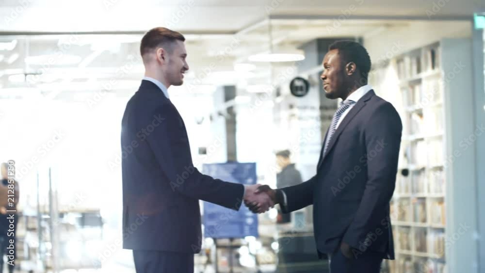 Young Caucasian businessman shaking hands and talking with African American colleague in the office Young Caucasian businessman shaking hands and talking with African American colleague in the office