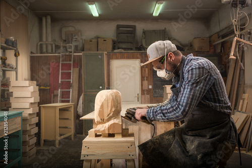 Сarpenter in work clothes and small buiness owner working in woodwork workshop, processes the board with an angle grinder , on the table is a hammer and many tools