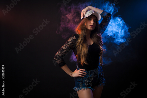 Young dark-haired woman in black top, jeans and cap posing against a background of blue smoke from a vape on a black isolated background