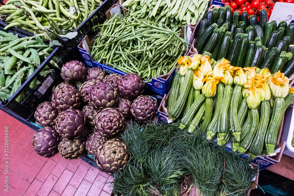 Market stall with fresh agretti, artichoke, courgette flowers, cucumber, tomatoes, fave and green beans with a purple floor background. No people. Rome. Europe.