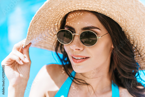 beautiful brunette woman in sunglasses and straw hat smiling at poolside