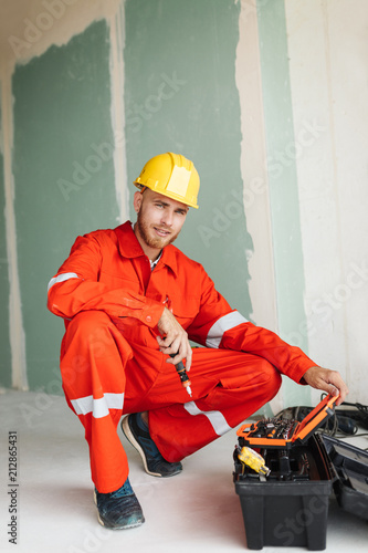Smiling foreman in orange work clothes and yellow hardhat dreamily looking in camera while opening chest with tools