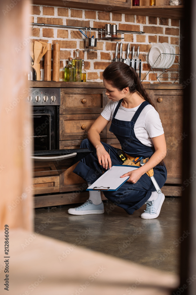 serious young repairwoman with clipboard sitting near oven and looking at it - obrazy, fototapety, plakaty 