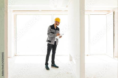 Young engineer in work clothes and yellow hardhat holding plan of apartments thoughtfully looking on wall with big window on background © Anton