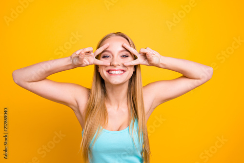 Portrait of young woman with blond hair, big blue eyes and toothy beaming smile. Happy girl showing v-sign with her two hands and looking at camera isolated on yellow background with copy space
