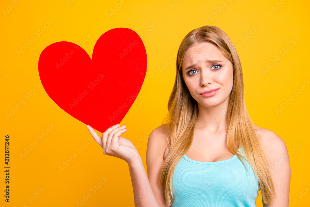 Portrait of sad and serious girl holding big paper carton heart figure. Young blond woman holding red heart in her hands isolated on yellow background Portrait of sad and serious girl holding big paper carton heart figure. Young blond woman holding red heart in her hands isolated on yellow background