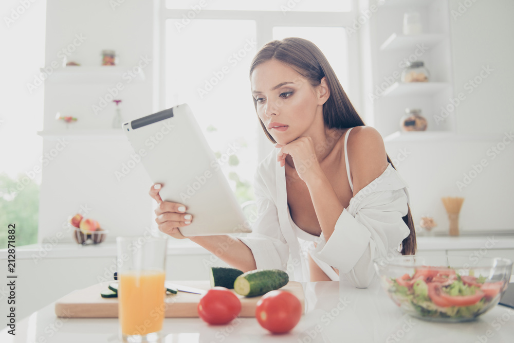 Sexy woman on white lingerie and long shirt with naked shoulder sit in thought, look at the tablet and biting lower lip. Kitchen with cutting board, ingredients for salad and juice on the table