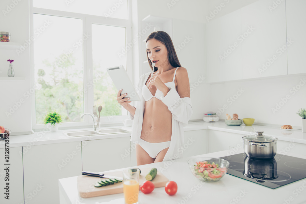 Hot brunette housewife with amazing body in white bra and panties look at the tablet thoughtfully holding fork in the mout. Girl reading recipe for a simple breakfast Hot brunette housewife with amazing body in white bra and panties look at the tablet thoughtfully holding fork in the mout. Girl reading recipe for a simple breakfast