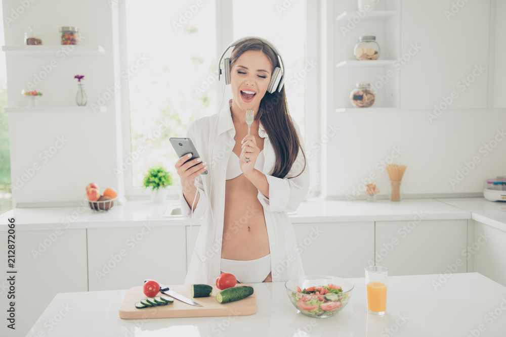 Good morning! Hot tempting and seductive woman in white lingerie and shirt with long sleeve, listening music through headphones and sings in the kitchen holds fork as a microphone Good morning! Hot tempting and seductive woman in white lingerie and shirt with long sleeve, listening music through headphones and sings in the kitchen holds fork as a microphone