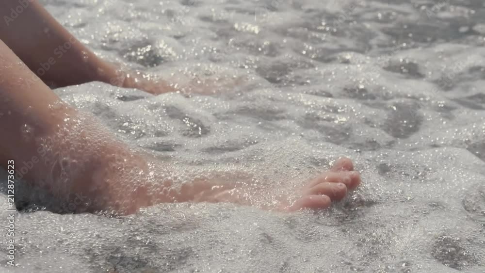 A close-up of a child's legs is overwhelmed by a wave. Pebble sea beach noisy sea waves on children's legs A close-up of a child's legs is overwhelmed by a wave. Pebble sea beach noisy sea waves on children's legs