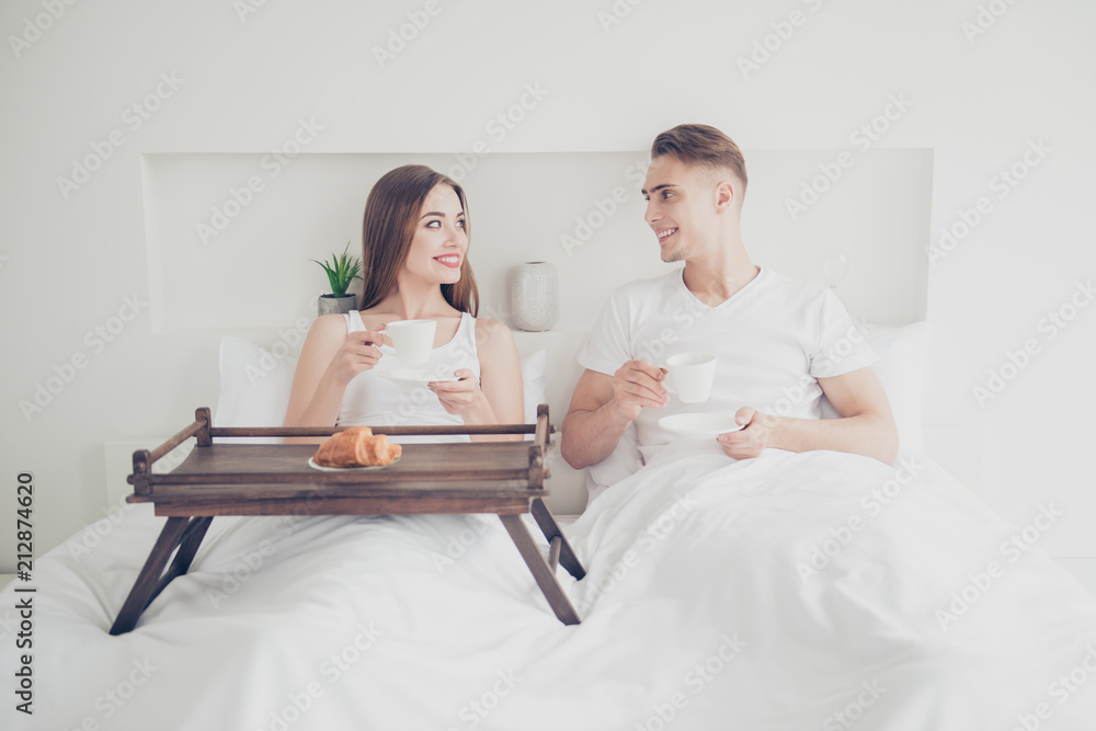 Portrait of young attractive cute couple sitting together in bed, covered with blanket, smiling, having breakfast, drinking coffee, eating croissants