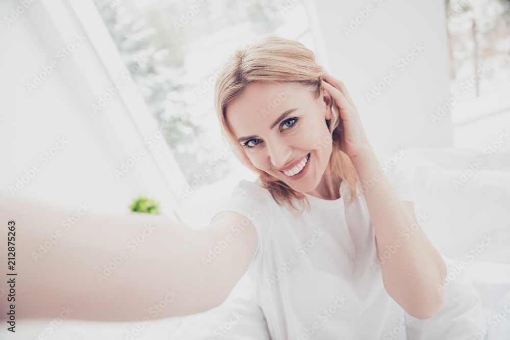 Self portrait of adult mature smiling blonde woman looking straight at camera touching her ear and hair with left hand on white interior background. Diet Self portrait of adult mature smiling blonde woman looking straight at camera touching her ear and hair with left hand on white interior background. Diet