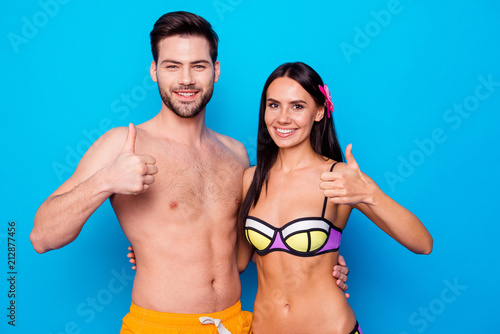 Positive joyful couple hugging wearing in swimsuit and show thumbs up sign. Concept of advertisement of summer vacations with woman with a woman with a flower in her hair and happy man