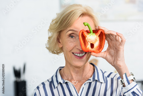 Foto attractive grey hair woman covering eye with half of bell pepper in kitchen and