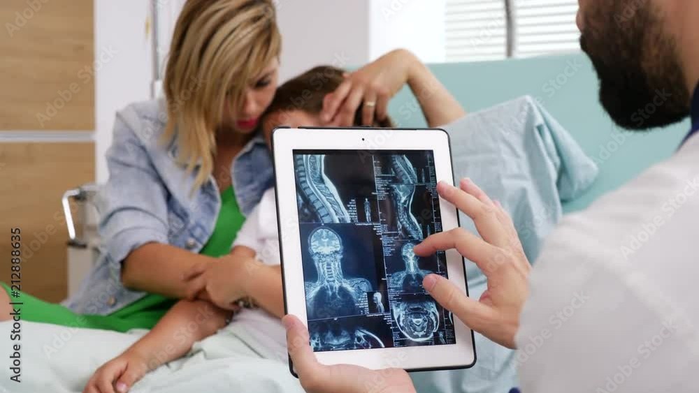 Doctor looking at MRI result on digital tablet PC screen while the girl patient and her mother are in the bed on blurred background Doctor looking at MRI result on digital tablet PC screen while the girl patient and her mother are in the bed on blurred background