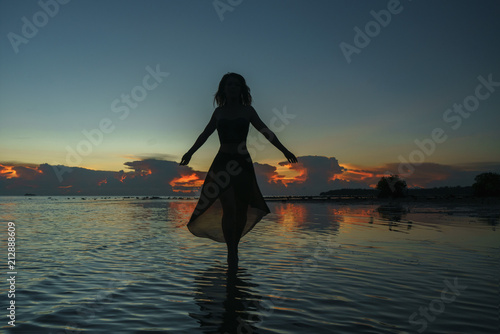 Happy young woman in a long dress dancing on the beach on a colourful sunset background