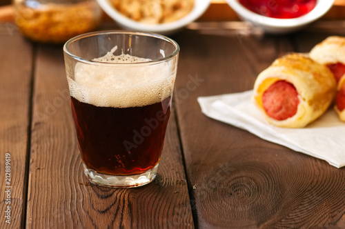 Close up of dark beer in a glass with pigs in a blanket, ketchup, nuts and mustard on background