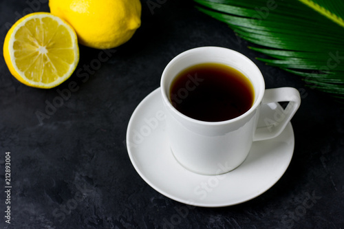 A cup of tea, a sliced lemon and a green leaf on a dark background