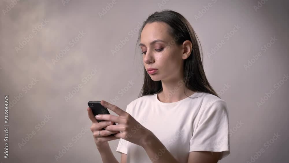 Young serious sexy brunette girl is typing message on smartphone, communication concept, grey background Young serious sexy brunette girl is typing message on smartphone, communication concept, grey background