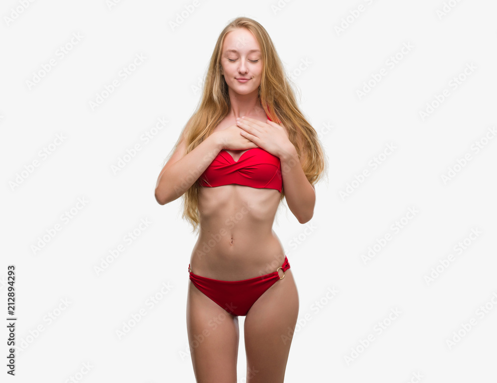 Obraz premium Blonde teenager woman wearing red bikini smiling with hands on chest with closed eyes and grateful gesture on face. Health concept.