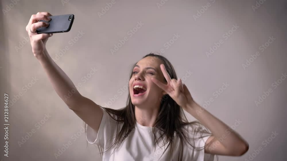 Young sexy brunette girl is making selfie on smartphone, fool around, watching at camera, smiling, communication concept, grey background Young sexy brunette girl is making selfie on smartphone, fool around, watching at camera, smiling, communication concept, grey background