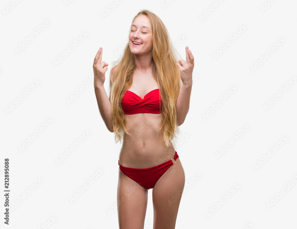 Blonde teenager woman wearing red bikini smiling crossing fingers with hope and eyes closed. Luck and superstitious concept. Blonde teenager woman wearing red bikini smiling crossing fingers with hope and eyes closed. Luck and superstitious concept.