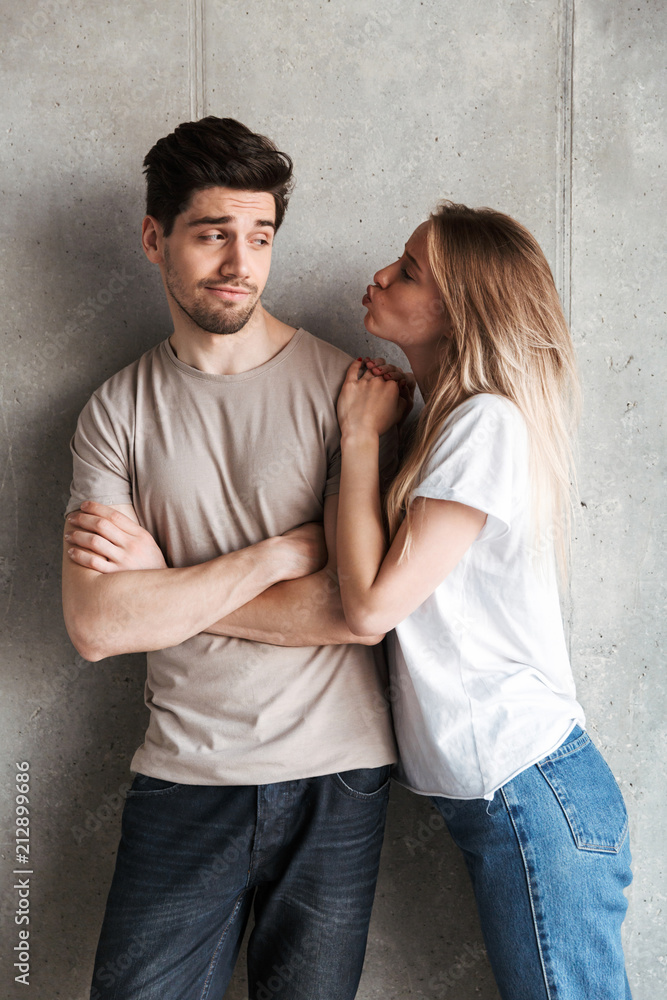 Portrait of young displeased man expressing unwillingness while beautiful woman kissing his cheek, isolated over gray background