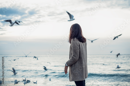 Woman in coat and trousers walking by seashore with flying seagulls