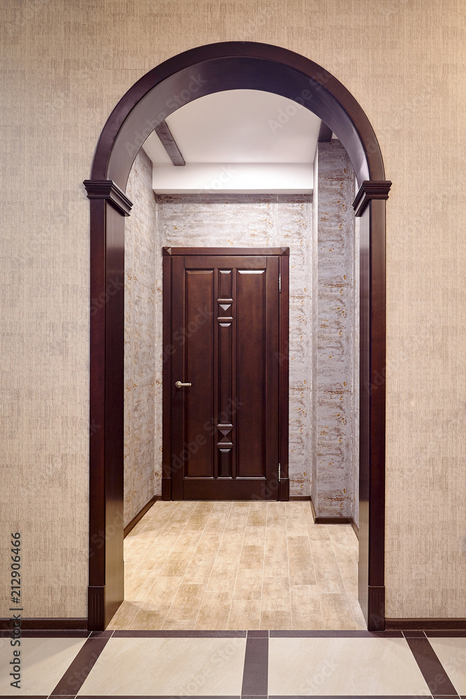 Fototapeta premium Passage through wooden archway into corridor with entrance door tiked in brown tones