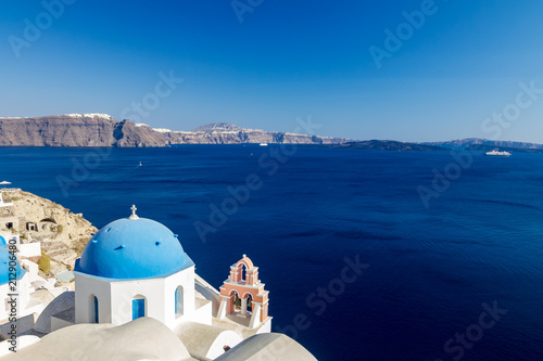 Incredibly romantic scene on Santorini. Fira, Greece. Amazing daytime view towards the deep sea crystal waters with white houses and blue church roof with white Christian cross and bells