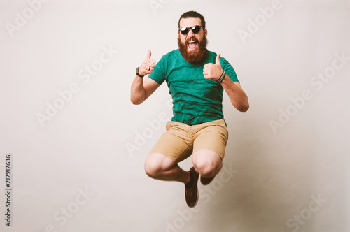 Fotografia Cheerful bearded hipster man with sunglasses jump over white background and show