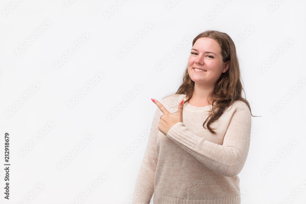 Happy caucasian young woman in sweater smiling broadly at camera, pointing fingers away, showing something interesting and exciting on studio wall. Copyspace for text Happy caucasian young woman in sweater smiling broadly at camera, pointing fingers away, showing something interesting and exciting on studio wall. Copyspace for text