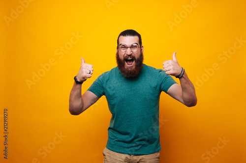 Amazed bearded hipster man wearing eyeglasses and showing thumbs up over yellow background