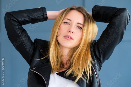 disheveled sexy young woman in black leather rocker jacket with ruffled hair. blond girl portrait on blue background