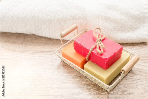 Three colorful soaps are placed in a basket, placed next to a towel on a wooden floor.