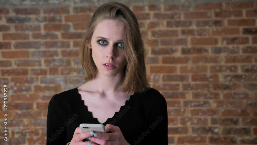 Young sexy girl with smoky eyes is typing message on smartphone, thinking process, communication concept, brick background Young sexy girl with smoky eyes is typing message on smartphone, thinking process, communication concept, brick background