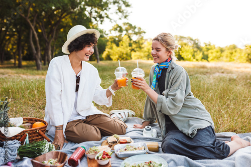 Pretty smiling girls sitting on picnic blanket holding orange juice in hands happily spending time on picnin in park