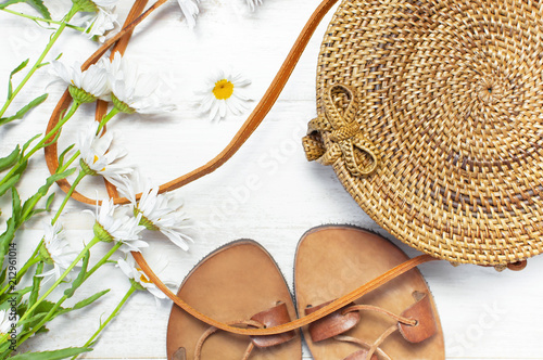 Fashionable handmade natural organic rattan bag, leather sandals, chamomile flowers on light wooden background. Ladies bag made of natural material. Copy space, top view. Ecobags from Bali.  photo