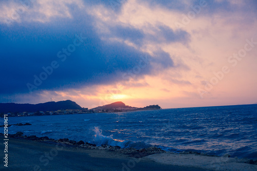Sunset Sea Perspective from the Shoreline, Beautiful Mountain and Ocean view Landscape, Shimmering Twilight with blue and dark colors, Hidding Sun between Cloudy Sky © Artur